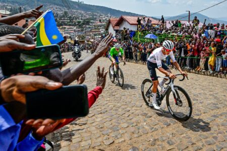 Cyclists race along a cobblestone street while a large crowd cheers from both sides, capturing the moment on phones and banners flagging the event behind them for a sporting event scene.