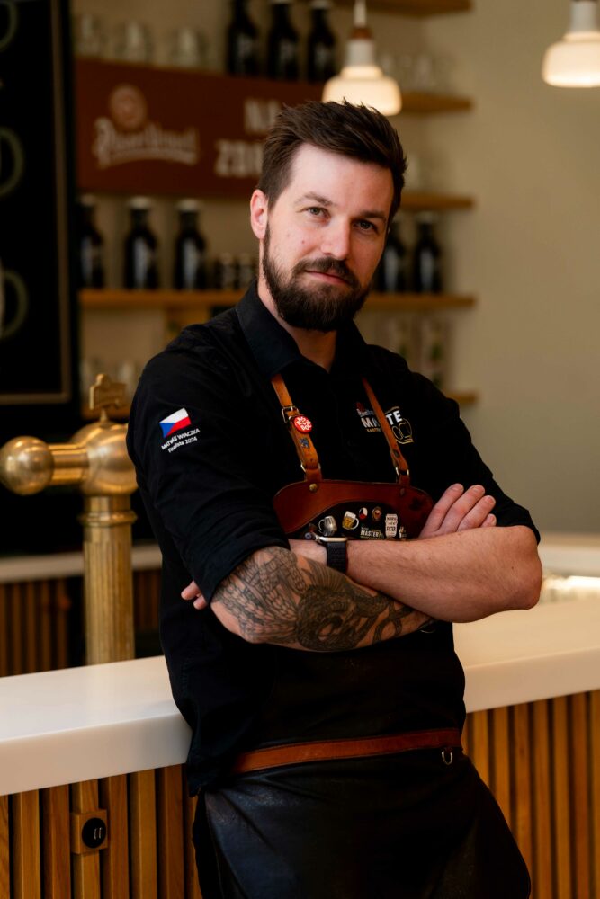 Portrait of a bearded man in a black shirt and leather apron, arms crossed, standing behind a bar/counter in a cafe or brewery setting.