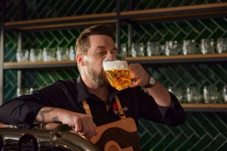Man with a beard in a black shirt and leather apron drinks a beer from a large mug at a bar, eyes closed.
