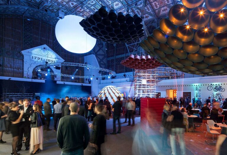 Crowd at a large indoor exhibition with futuristic hanging sculptures and a bright circular orb overhead.