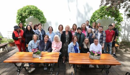 Outdoor group photo of about 25 adults posing together in a courtyard with wooden benches and leafy arches above them; sunny day.
