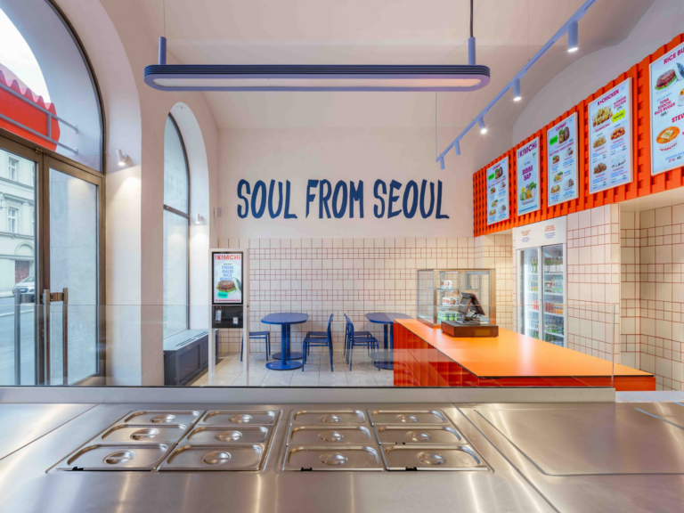 Bright interior of a Seoul-themed fast-casual restaurant with a blue 'SOUL FROM SEOUL' mural, orange counter, and menu boards on the wall behind the counter.