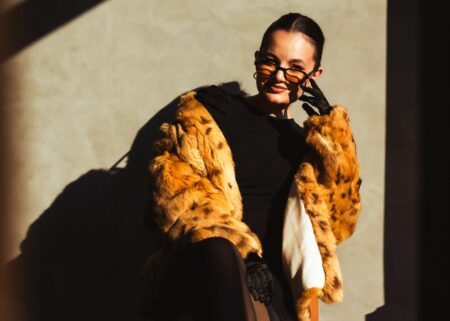 Woman in a black dress and leopard fur coat sits against a sunlit beige wall, adjusting orange sunglasses.