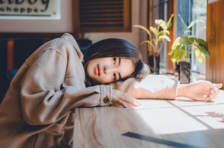 Woman in a beige coat rests her head and arm on a sunlit cafe table by a window.