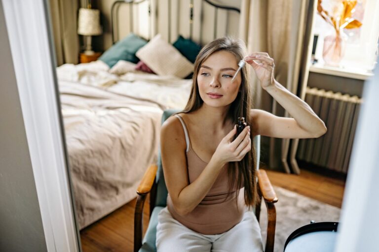 Woman applying skincare serum to her face with a dropper, seated in a bedroom with a bed and pillows in the background.