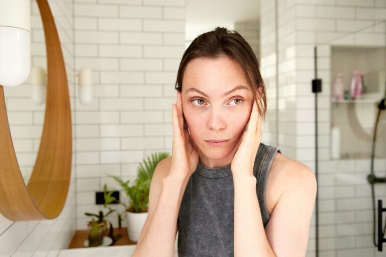 Woman in a grey sleeveless top cupping her face and looking worried in a tiled bathroom mirror area