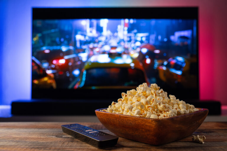 Dřevěná miska s popcornem a dálkový ovladač před televizí při domácím sledování filmu nebo seriálu. Zdroj fotografie: Adobe Stock.