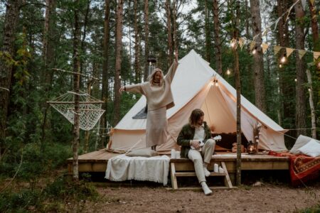Romantický glamping v lese – pár odpočívá před stylovým stanem osvětleným světýlky v přírodě. Zdroj fotky: Pexels